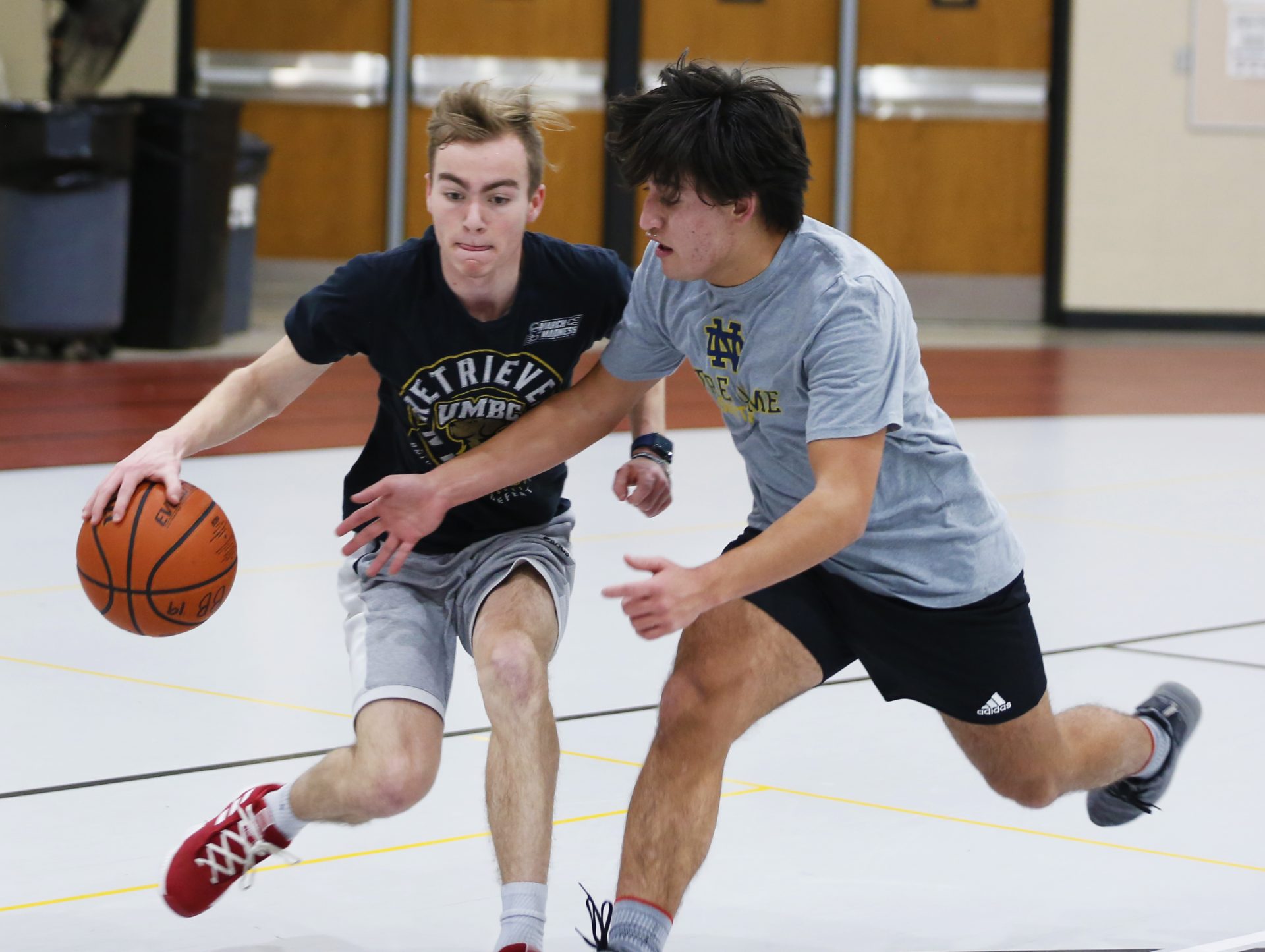 Two male basketball players in a gymnasium. One player is holding a basketball while running towards the other player.