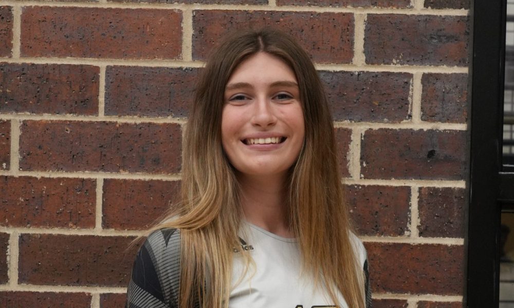Person standing in front of brick wall, smiling at camera, wearing a white shirt with number twelve and black shorts, posing