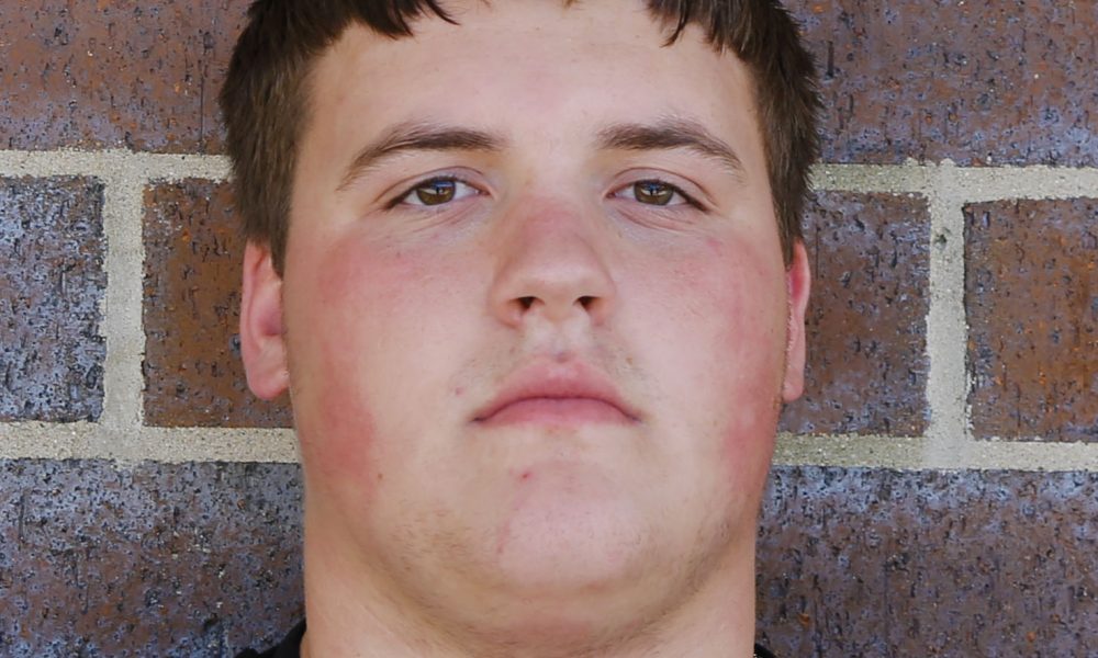Young man with short brown hair, wearing a black shirt against a brick wall background.