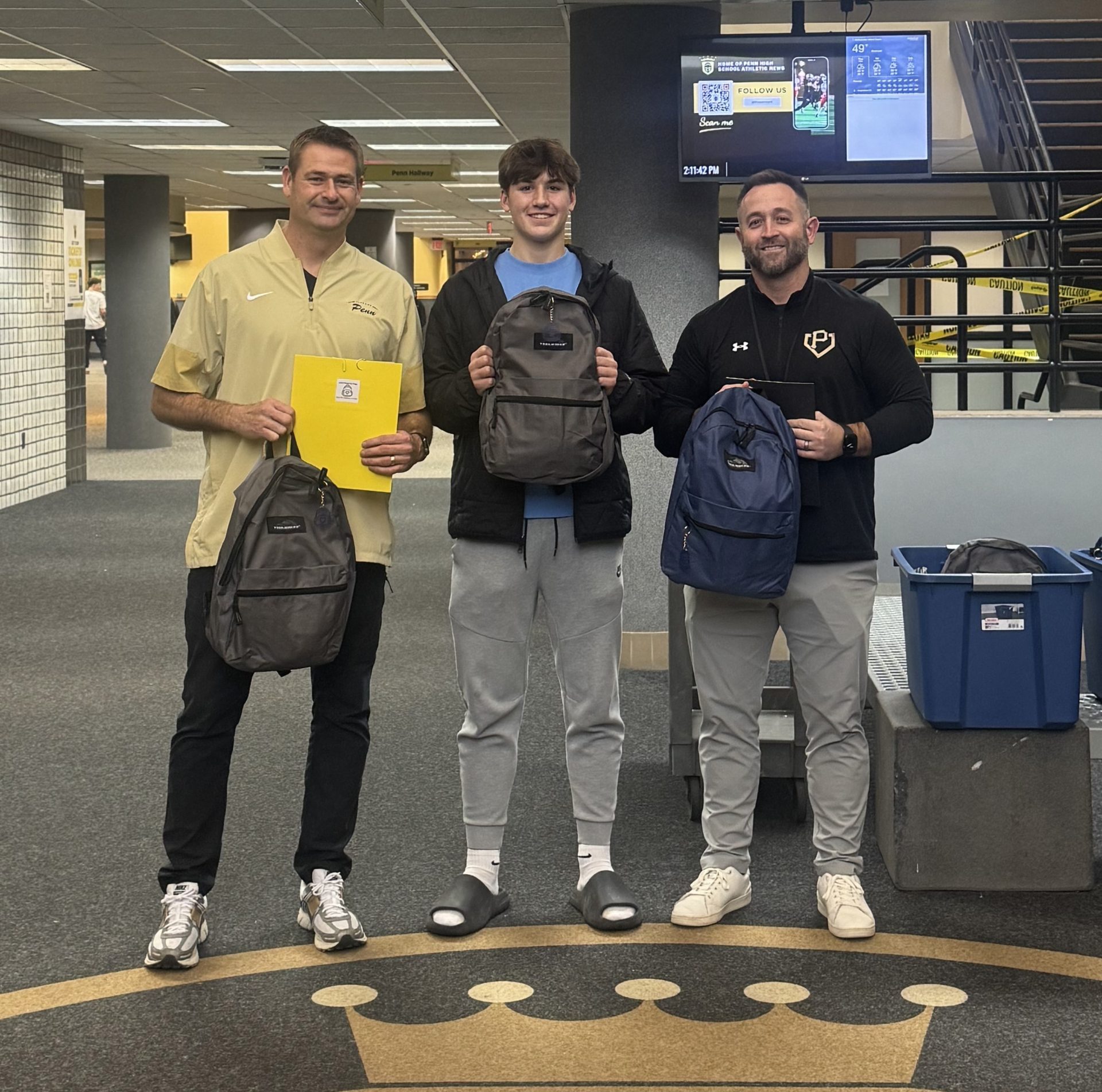 From left, Penn Football Head Coach Pete Riordan, Will Abercrombie and Counseling Center Leader John Westra display Little Blessing Backpack Bags in the commons area of Penn High School.