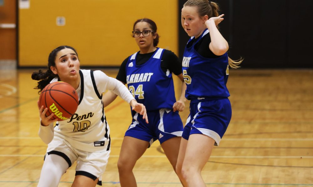 Three girls playing basketball on a court. One girl is shooting the ball while two others are defending. The player in the
