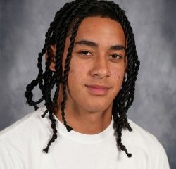 Young male with dreadlocks wearing a white t-shirt and braided hair, posing for school portrait.