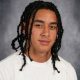 Young male with dreadlocks wearing a white t-shirt and braided hair, posing for school portrait.