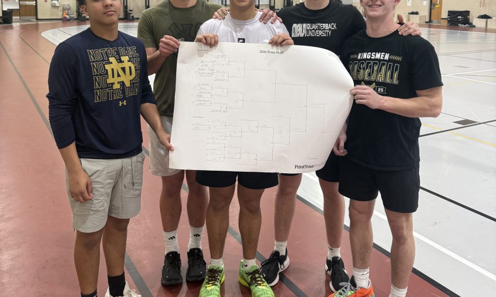 Five young men posing for a group photo in front of a basketball court, each wearing different sports attire and holding a