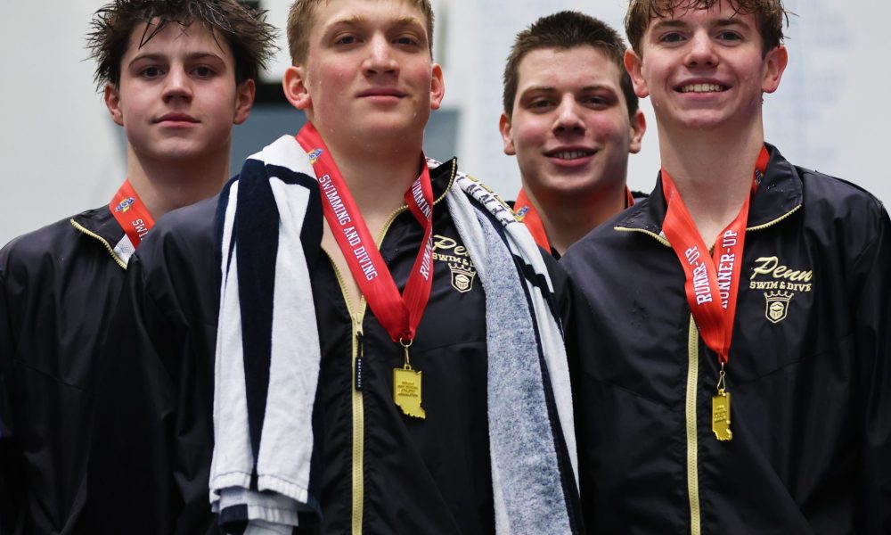 Five young male athletes posing in front of a sign, each wearing medals around their necks and holding towels.