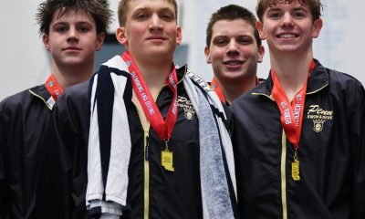 Five young male athletes posing in front of a sign, each wearing medals around their necks and holding towels.