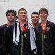 Five young male athletes posing in front of a sign, each wearing medals around their necks and holding towels.