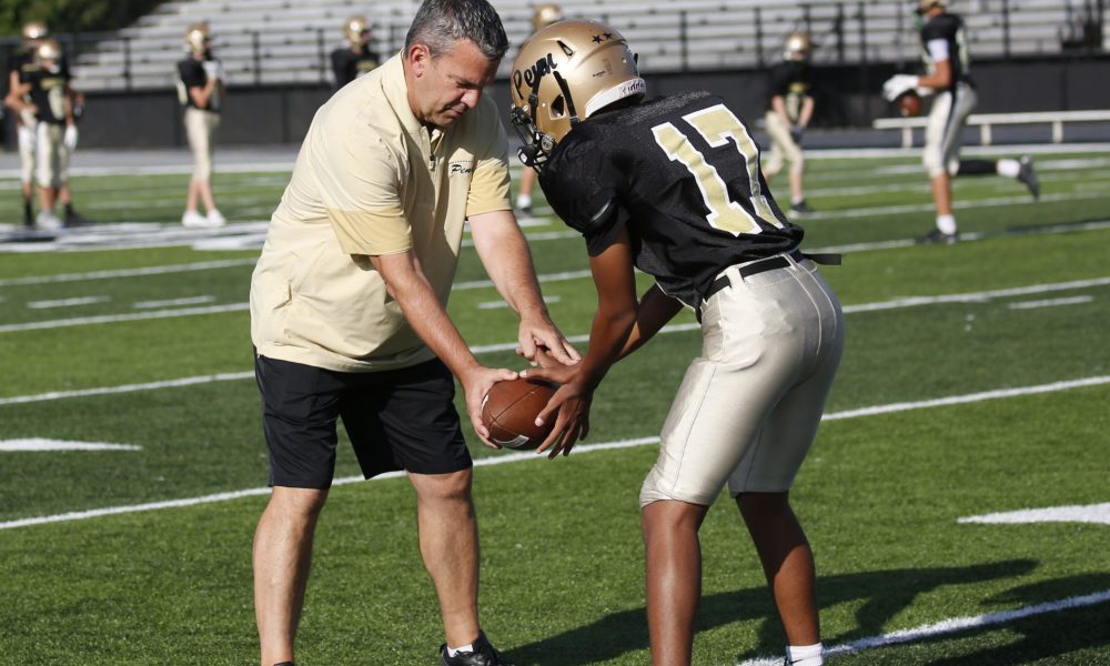 Coach O'Connell on the football field during practice showing a player how to hold a football.