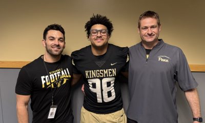 Three men posing together in front of a wall. The man on the left is wearing a football uniform and holding a football. The