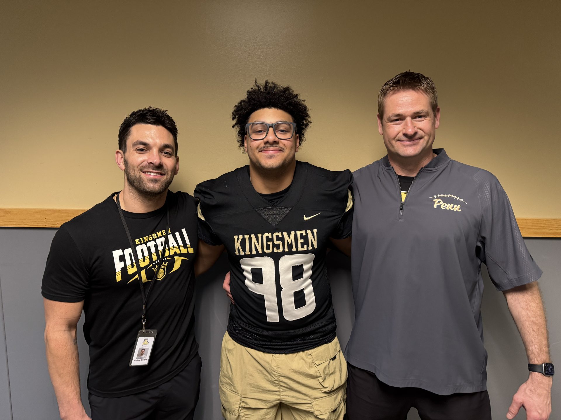 Three men posing together in front of a wall. The man on the left is wearing a football uniform and holding a football. The