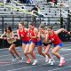 Female runners in a race, all wearing red uniforms, sprinting on a track.