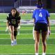 Two women on a football field, one in a black uniform and the other in a blue uniform, both wearing cleats